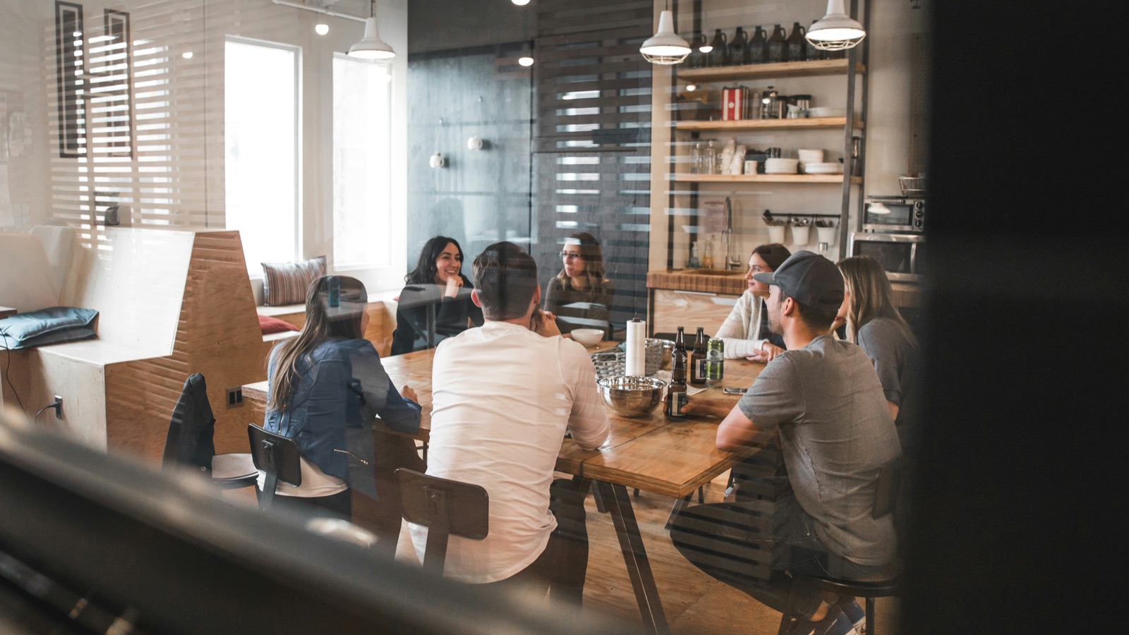 Seven people talking around a long wooden table in an office kitchen